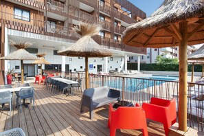 A wooden deck with outdoor seating, a pool, and a building with balconies in the background. - Appartement 5 Minutes de la Plage | Accès Sauna (Sète)