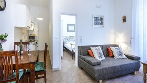 A modern living room with a grey sofa, wooden dining table, and chairs. A kitchen with white cabinets and a microwave is visible in the background. - Apartments Franka - One-Bedroom Apartment (Dubrovnik)