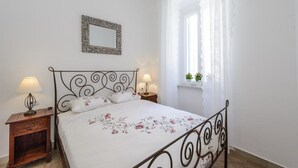 A bedroom with a bed, two bedside tables, a nightstand, a window with curtains, and a wall-mounted mirror. - Apartments Franka - One-Bedroom Apartment (Dubrovnik)