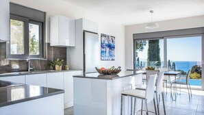 A modern kitchen with a central island, white cabinetry, and a large window overlooking the sea. - Villa Della Luna - Three Bedroom Villa, Sleeps 6 (Vlachata)