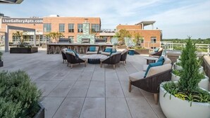 Un espace extérieur aménagé en terrasse, avec des sièges et un bar, entouré de plantes en pot et d’un bâtiment sur lequel est apposée une enseigne indiquant « LiveGalleryApts.com ».