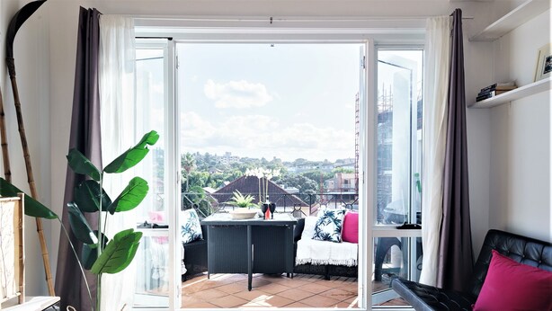 Una sala de estar moderna con un ventanal corredera que da a un balcón, un sofá negro con un cojín rojo y una mesa de mimbre con una planta.