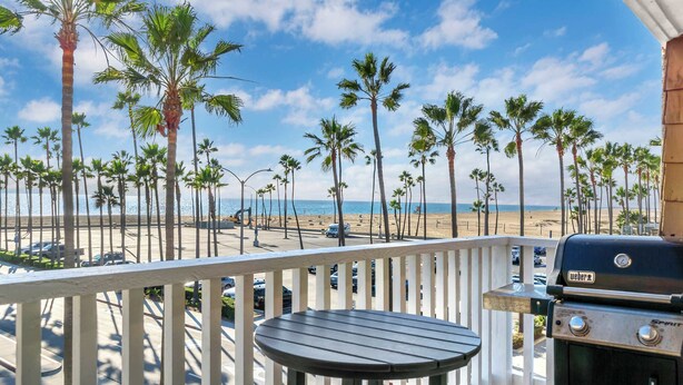 Un balcón con una parrilla y mesa con vista a una playa con palmeras y cielo despejado.