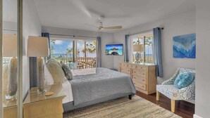 Un dormitorio con cama, cómoda, silla, ventilador de techo y un amplio ventanal con vista a palmeras y una playa.