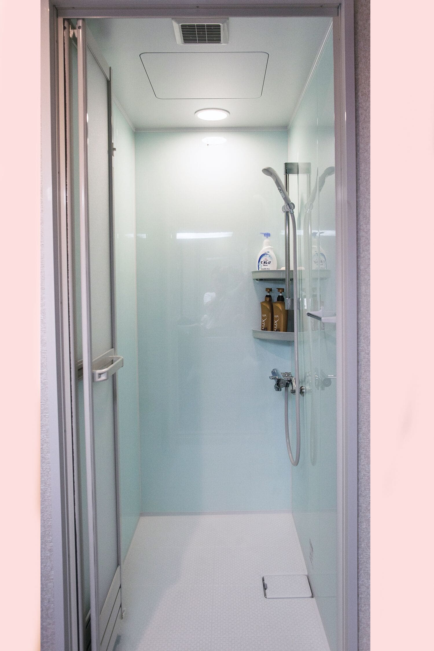 A glass-enclosed shower stall with a white floor, a glass door, and a shelf with toiletries.