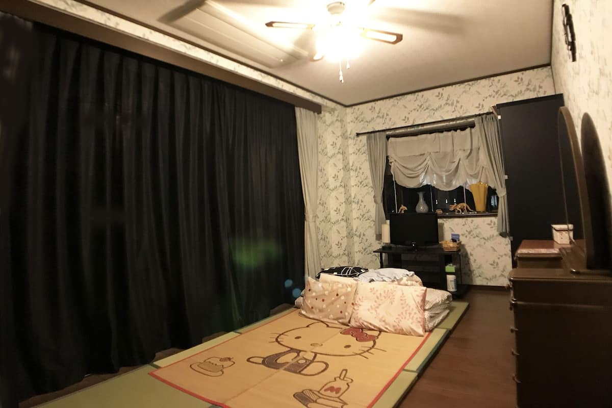A room with a futon bed, a small table, a cabinet, and a window with curtains.