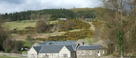 A traditional stone house with a sloped roof, surrounded by a fenced property and set against a backdrop of rolling hills and trees.