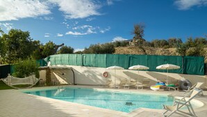Una piscina con sillas de descanso y sombrillas, rodeada por una cerca verde y árboles.