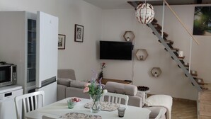 Uma sala de estar moderna com um sofá branco, uma mesa de jantar com um vaso de flores, uma televisão de tela plana na parede e uma escadaria com um mural tropical.