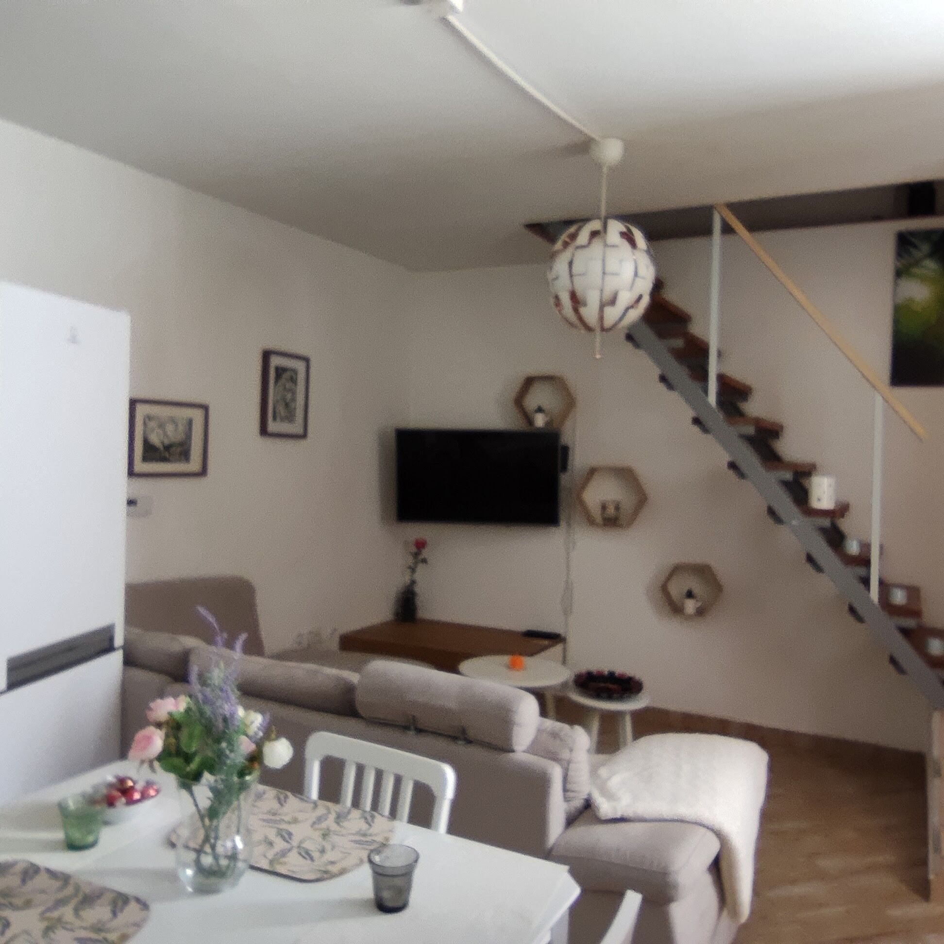 A living room with a sofa, a dining table with chairs, a TV mounted on the wall, and a staircase.