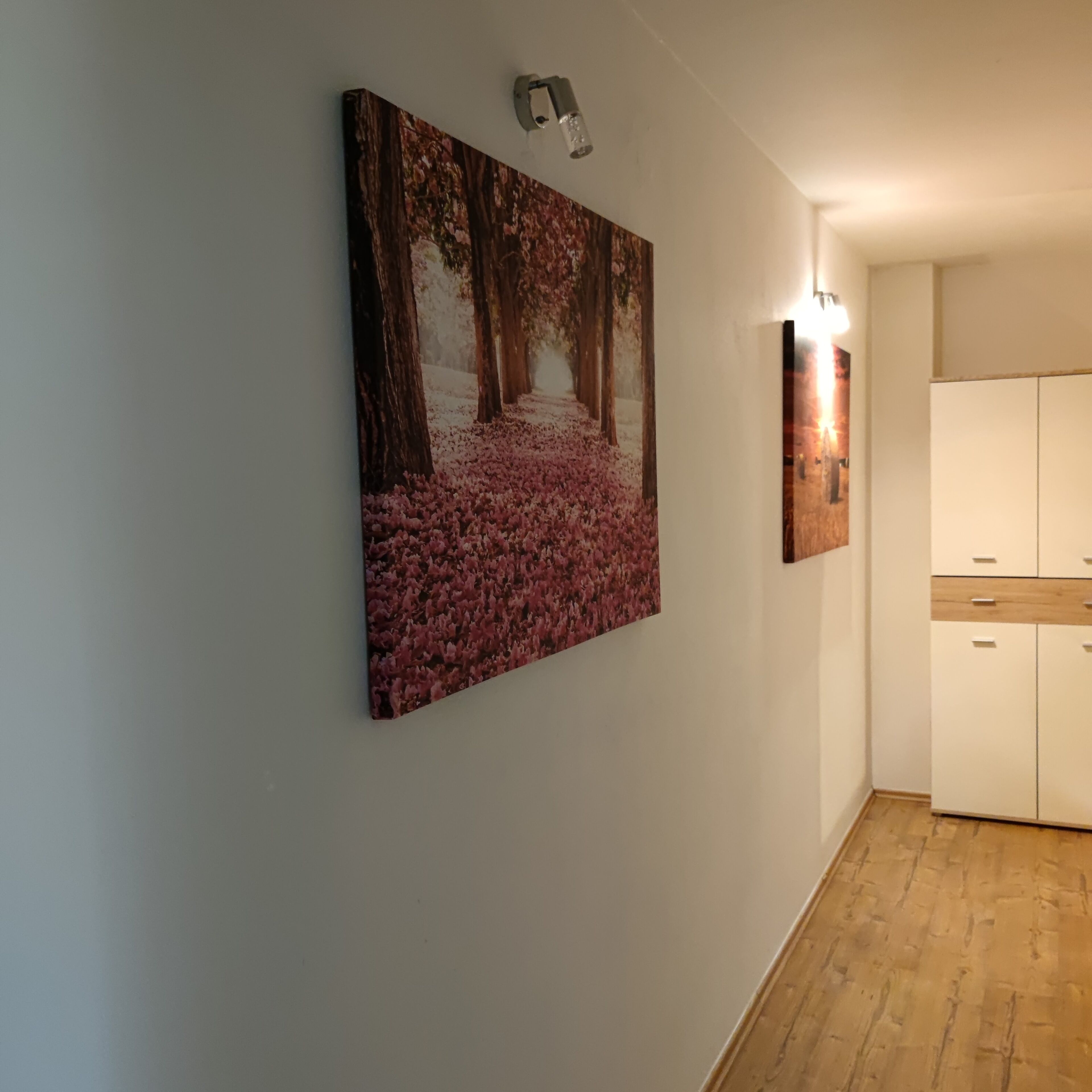 A hallway with a large painting of a forest scene on the wall, a light fixture, and a wooden cabinet.