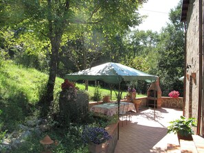 An outdoor patio with a table and chairs under a green umbrella, surrounded by lush greenery and a stone oven.