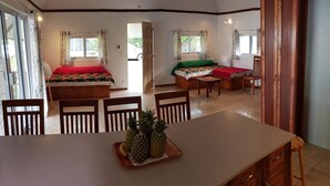A spacious room with a large dining table, two beds with colorful bedding, wooden cabinets, and a sliding glass door leading outside. - FARE AT MARIE LOU (Papetoai Moorea)