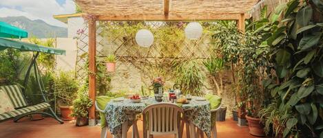 A covered patio with a table set for two, surrounded by potted plants and a bamboo trellis.