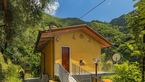 A yellow building with a wooden door and a balcony, surrounded by greenery and hills.