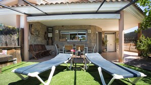 A covered patio with two lounge chairs, a table with a bottle, and a stone wall.