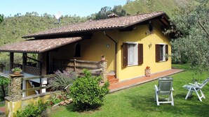 Una villa amarilla con techo de tejas, una escalera de piedra y un pequeño jardín con plantas y césped.