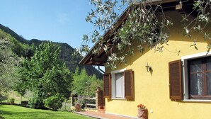 Un edificio amarillo con contraventanas de madera, una escalera de piedra y un jardín con césped y árboles.