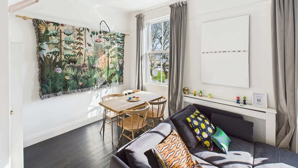 Un salon chaleureux avec un canapé foncé, une table à manger avec des chaises, une grande tapisserie et une fenêtre avec des rideaux.