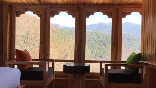 Habitación de cabaña de madera con cama, dos sillas, una mesita y ventanas amplias con vistas a la montaña.