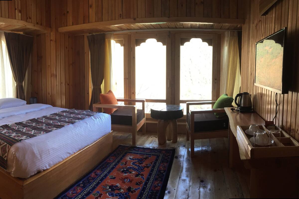 A wooden cabin room with a bed, a desk, a chair, a table, and a window with curtains.