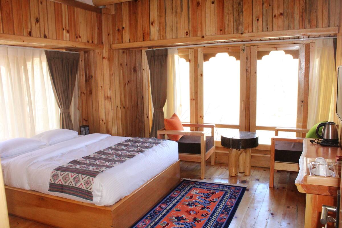 A wooden room with a bed, two chairs, a small table, and a window with curtains.