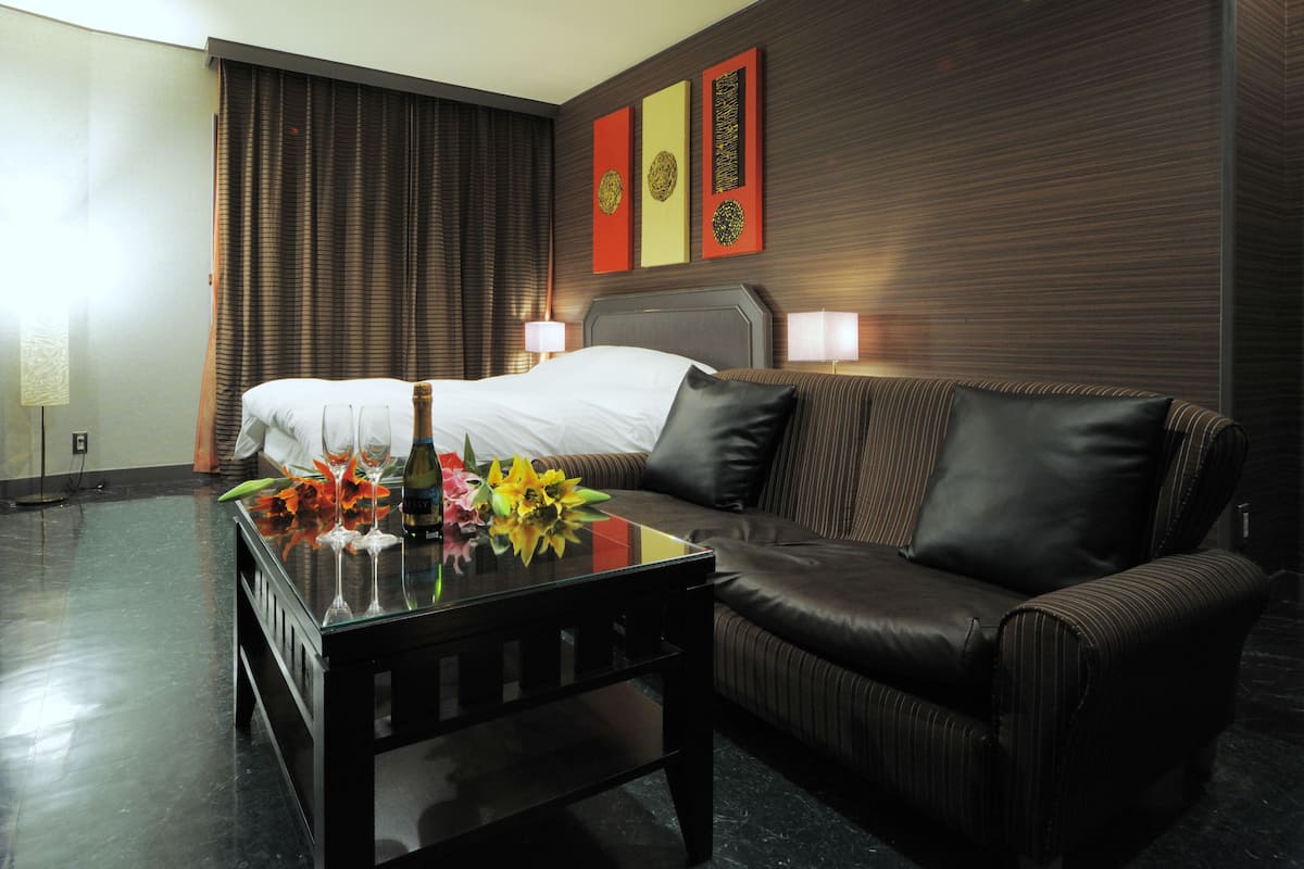 A hotel room with a bed, a leather sofa, a glass coffee table with flowers, and wall art.