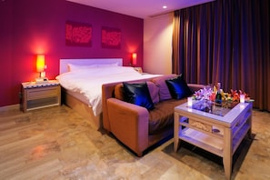 A hotel room with a large bed, a sofa, a small table with a bottle and flowers, and two nightstands with lamps.