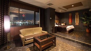 A hotel room with a large window offering a city view, a sofa, a bed, a coffee table, and a potted plant. - Hotel Sol - Adults Only (Fukuoka)