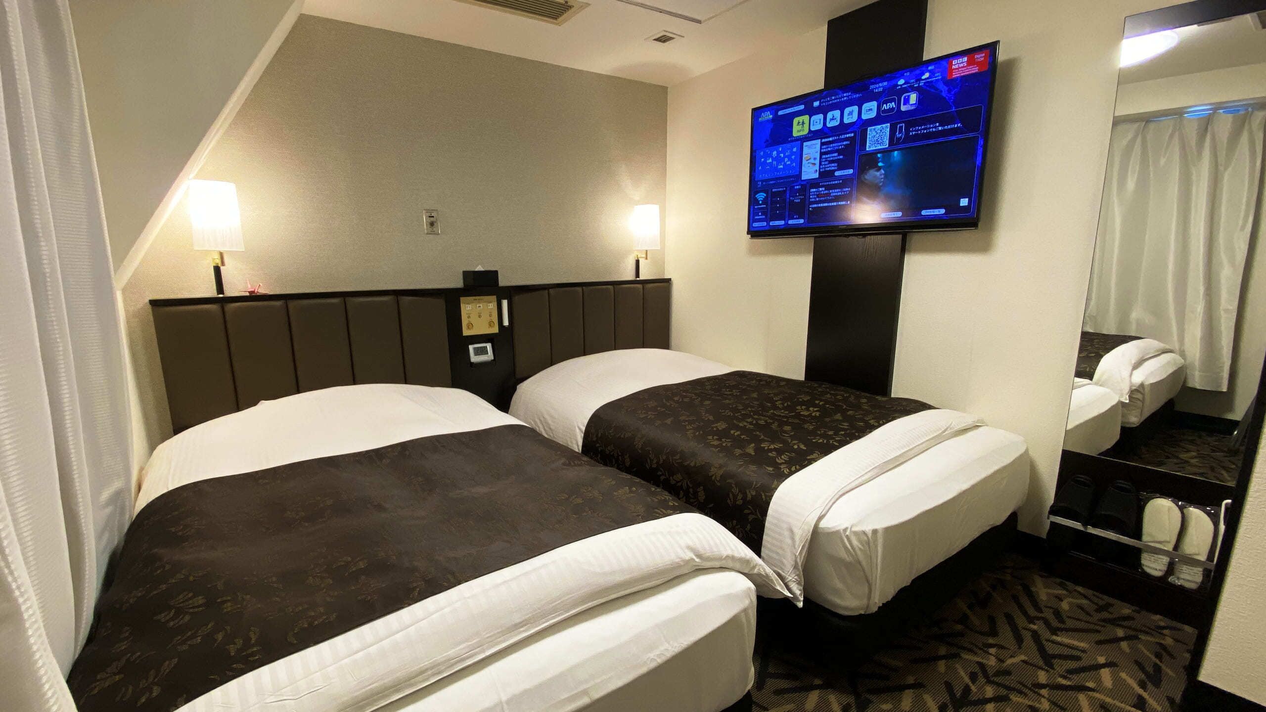 Une chambre d’hôtel avec deux lits, un téléviseur à écran plat fixé au mur, une table de chevet et un miroir.