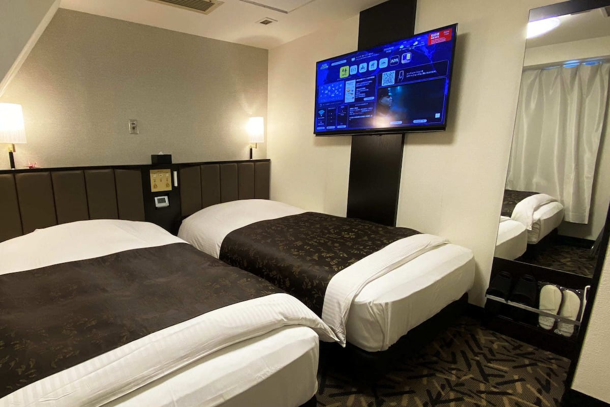 A hotel room with two beds, a flat-screen TV mounted on the wall, a bedside table, and a mirror.