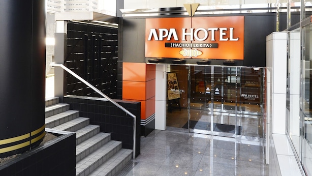 The entrance to APA HOTEL with a modern design, featuring a staircase leading to the entrance, a glass facade, and a prominent sign above the door.
