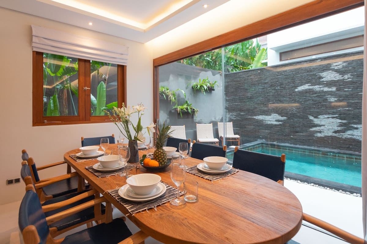 Un comedor con una mesa redonda de madera dispuesta para cuatro personas, rodeada de sillas. Hay platos, tazones y vasos sobre la mesa, además de un florero con flores. La habitación tiene ventanas amplias, vista a una piscina y una pared con plantas.
