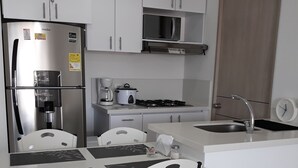 A modern kitchen with white cabinets, a stainless steel refrigerator, a gas stove, and a sink. There are three white chairs with black frames and a white table. - Comfort, NATURE, REST AND FUN WITH THE FAMILY (RICAURTE CUNDINAMARCA)