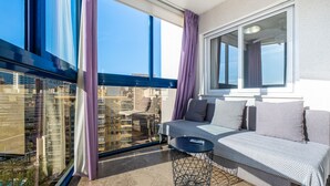 A balcony with a sofa, a small round table, and a view of a cityscape through large windows. - Acintur 13-A (Benidorm)