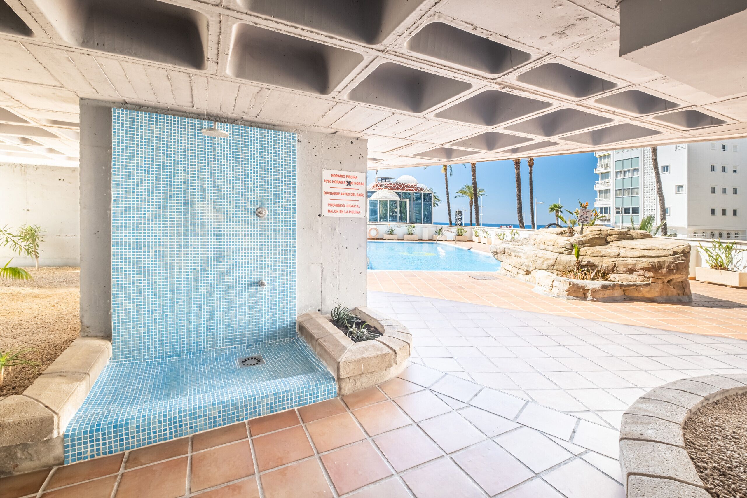 A swimming pool area with a tiled wall, a sign indicating pool hours, and a view of a building in the background.