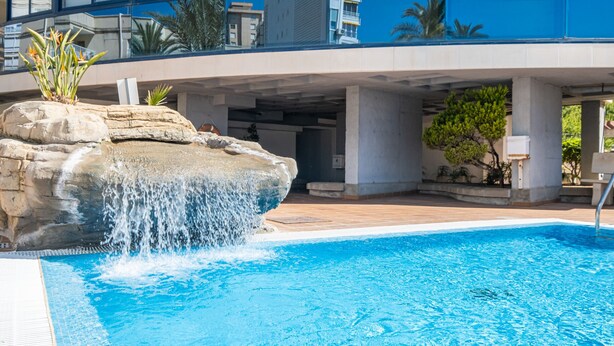A swimming pool with a waterfall feature, surrounded by a concrete deck and some greenery. - Acintur 13-A (Benidorm)
