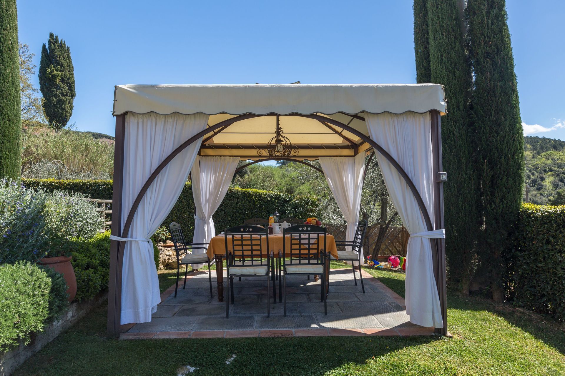 A covered outdoor dining area with a table and chairs, surrounded by a garden with hedges and trees.