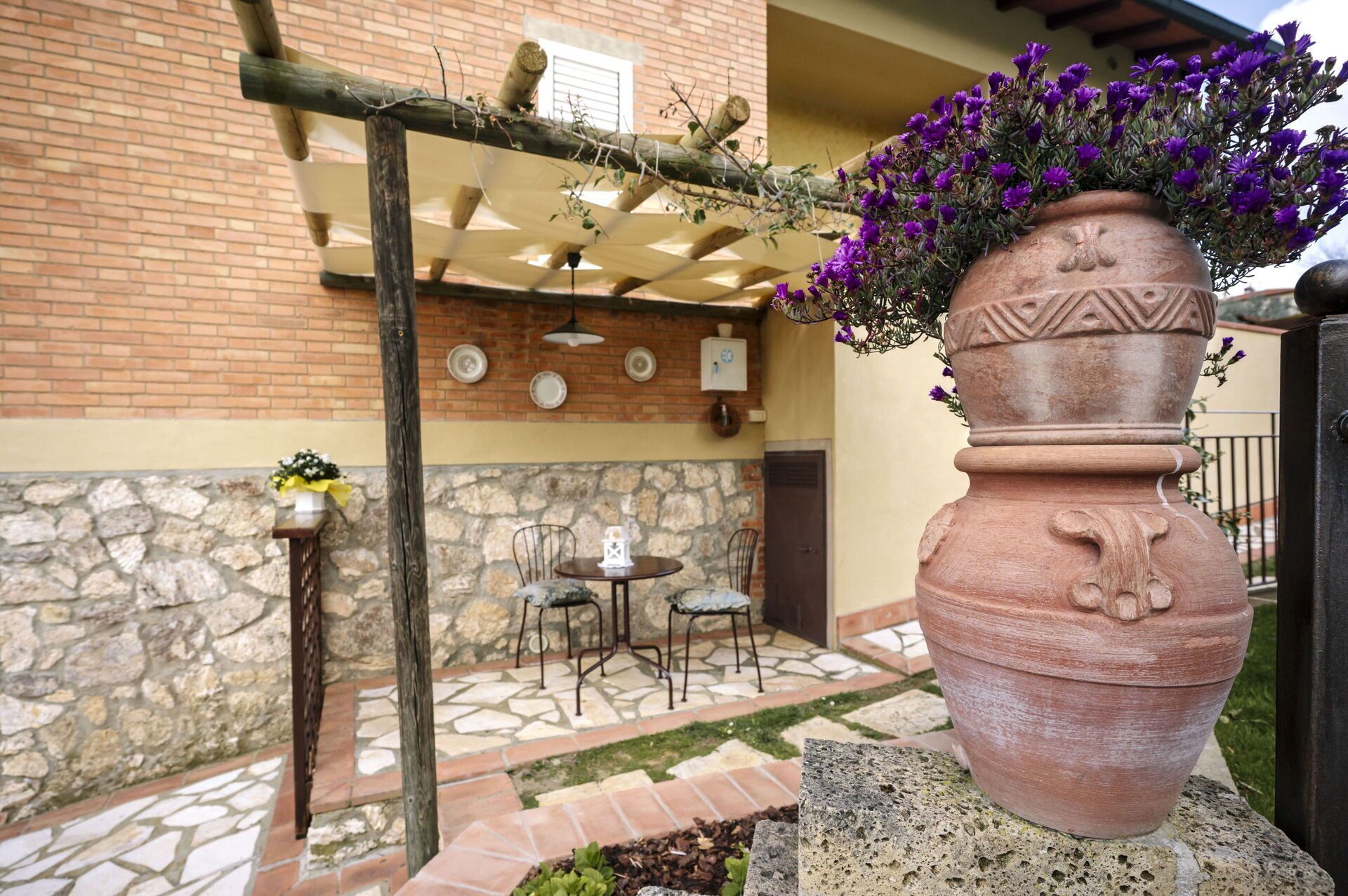 En patio med murstensvæg, stenbelægning og en stor terracotta krukke med lilla blomster.