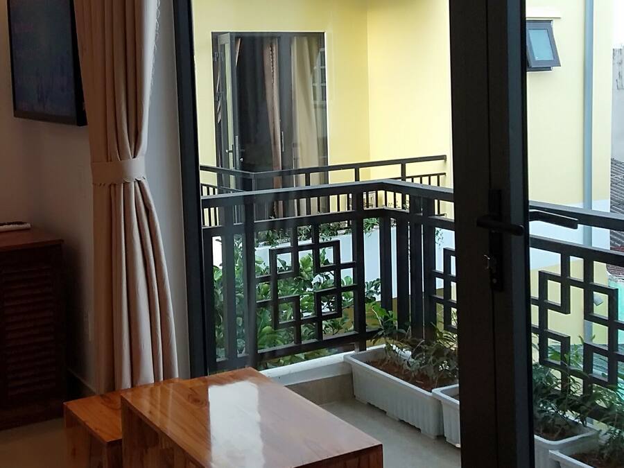 A modern living room with a wooden coffee table and a sofa, a large sliding glass door leading to a balcony with a black metal railing, and a view of a yellow building outside.