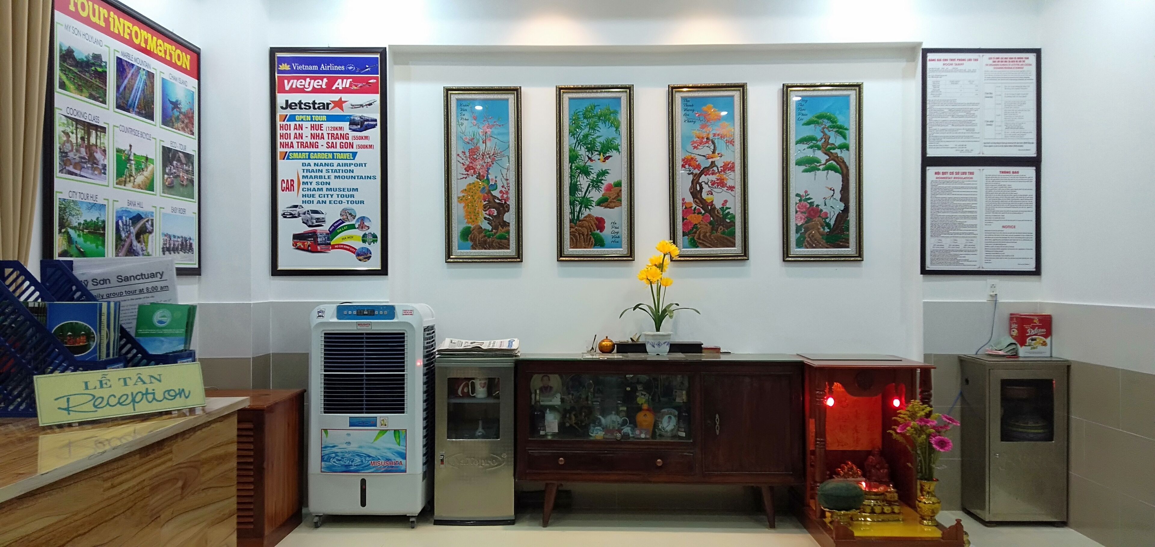 A reception area with a sign reading "LE TAN Reception," a large poster with travel information, and three framed paintings on the wall.