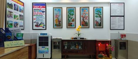 A reception area with a sign reading "LE TAN Reception," a large poster with travel information, and three framed paintings on the wall.