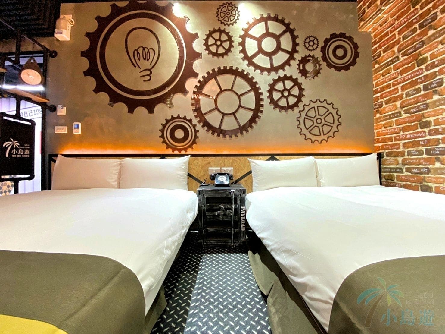 A hotel room with two beds, a wall decorated with gears and a light bulb, a brick wall, and a small table with a clock.