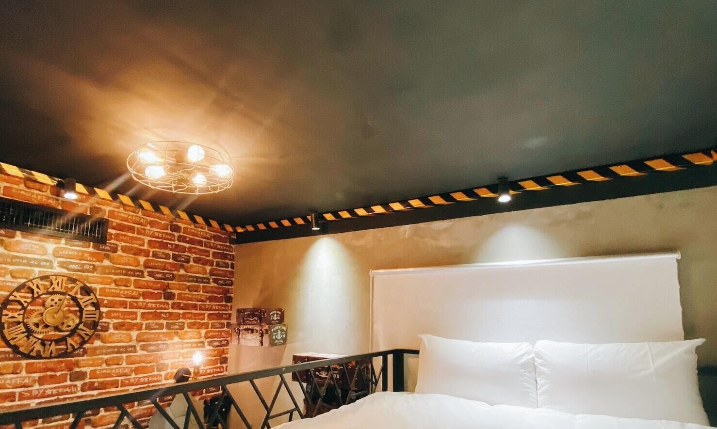 A room with a brick wall, a bed with white bedding, a round clock, and a ceiling light fixture.
