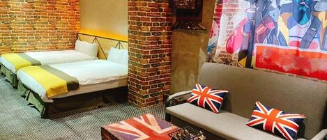 Una habitación con pared de ladrillo, dos camas, un sofá y una mesa con un cojín con la bandera Union Jack.