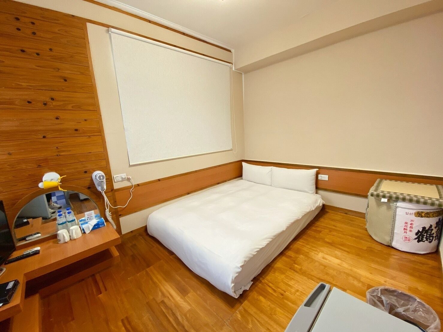 A hotel room with a wooden floor, a bed with white bedding, a desk with a phone, a mirror, and a small table with toiletries.