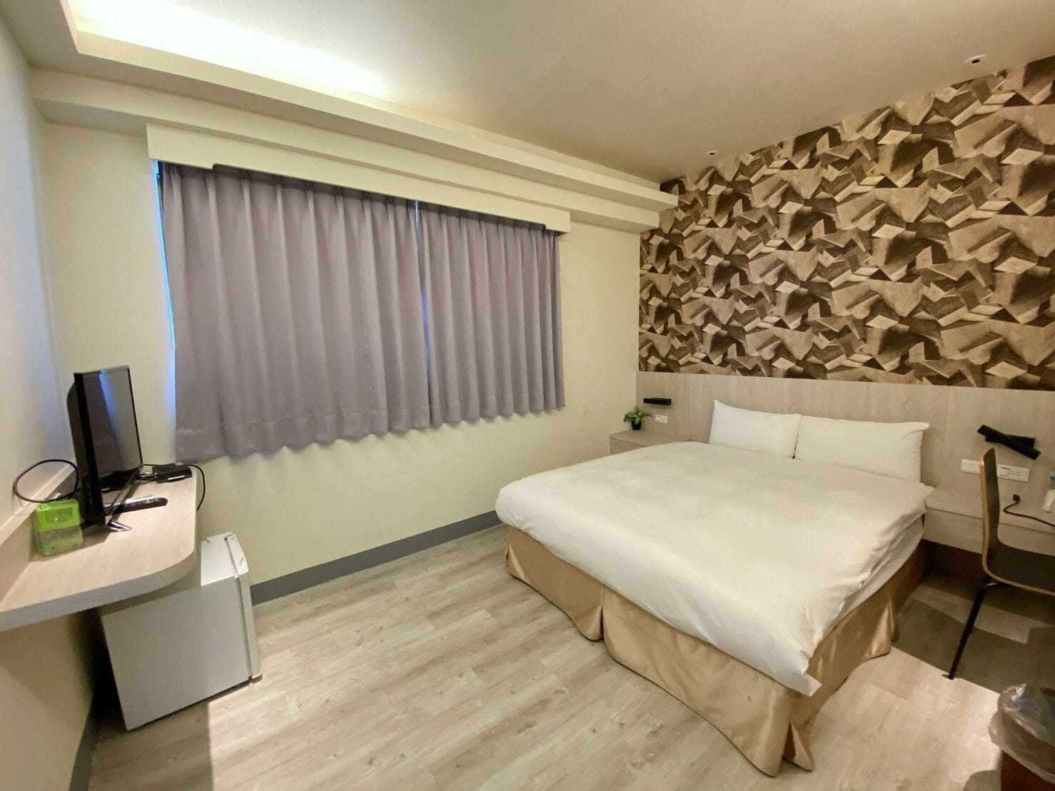 A hotel room with a bed, a desk with a computer, a small fridge, and a wall with a geometric pattern.