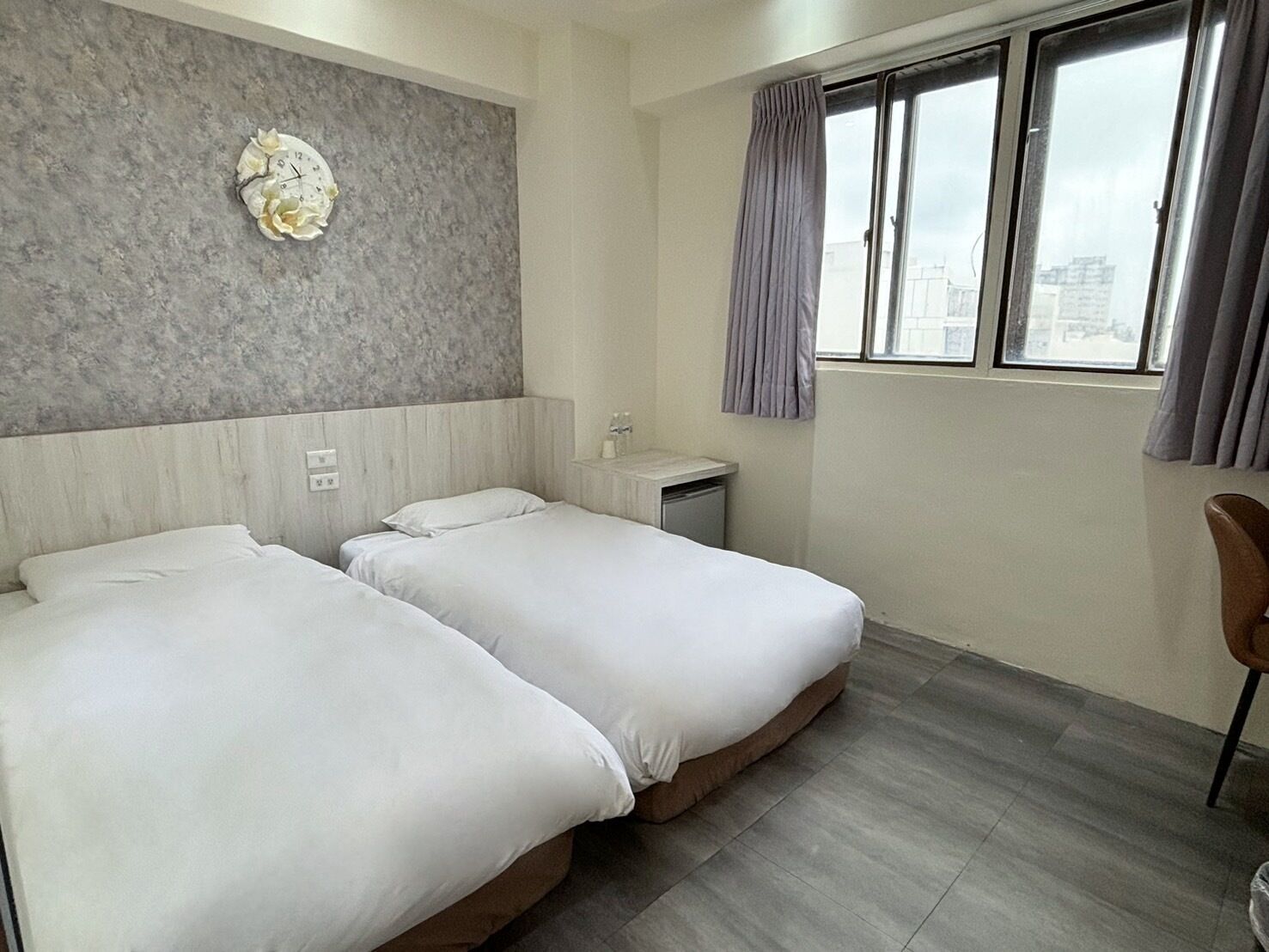 Habitación de hotel con dos camas, una pared gris con textura, una ventana con cortinas y vista a la ciudad.