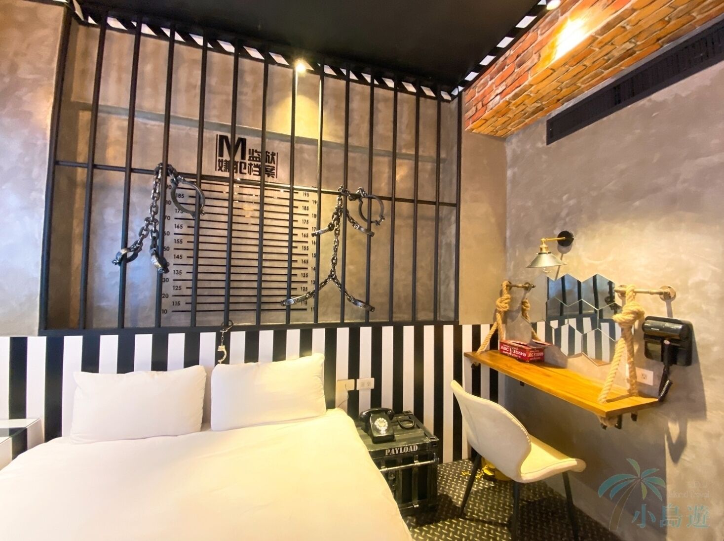 Ein Hotelzimmer mit einem Bett, einem Schreibtisch, einem Stuhl, einem kleinen Tisch mit einer Lampe sowie einer Wand mit einer Größenangabe und Ketten.