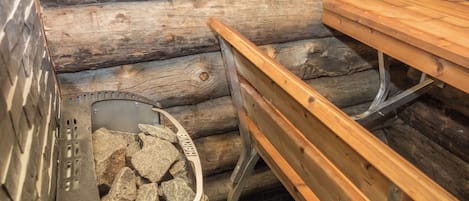 Un banco de madera y una parrilla metálica con piedras en su interior, frente a un fondo de paredes de troncos.